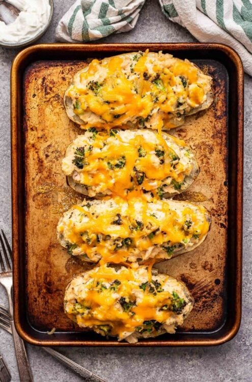 Four Broccoli cheddar chicken twice baked potatoes on a sheet pan. A dish towel and bowl of sour cream behind the pan.